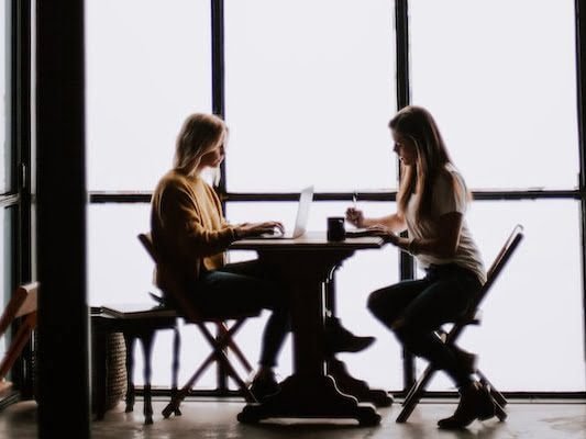 photo of two women facing each other sitting in front of table near glass wall